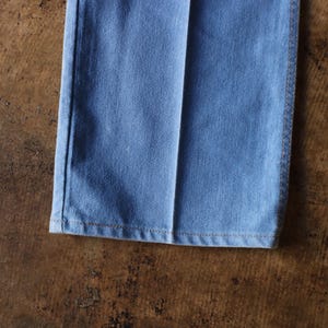 May include: Close-up of the bottom of light blue denim pants. The hem is visible, with a seam running along the edge. The pants are on a brown surface.