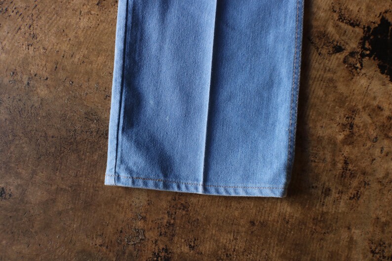 May include: Close-up of the bottom of light blue denim pants. The hem is visible, with a seam running along the edge. The pants are on a brown surface.