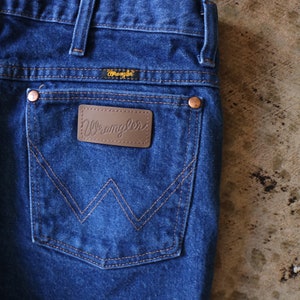 May include: Dark blue denim jeans featuring a back pocket with a brown leather patch embossed with the Wrangler logo. The pocket displays a signature W-shaped stitching pattern in orange thread, copper-colored rivets, and a small Wrangler label.
