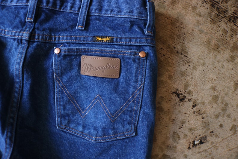 May include: Dark blue denim jeans featuring a back pocket with a brown leather patch embossed with the Wrangler logo. The pocket displays a signature W-shaped stitching pattern in orange thread, copper-colored rivets, and a small Wrangler label.
