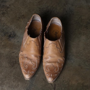 May include: A pair of light brown leather ankle boots with pointed toes. The boots have decorative cut-out patterns on the front and sides. The shoes are set against a dark, textured background.