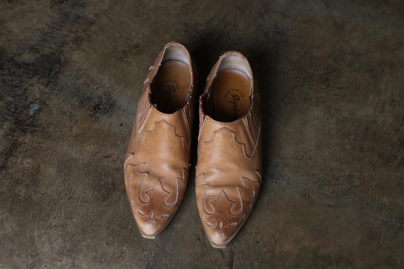 May include: A pair of light brown leather ankle boots with pointed toes. The boots have decorative cut-out patterns on the front and sides. The shoes are set against a dark, textured background.