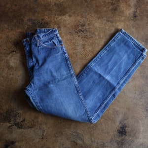 May include: A pair of blue denim jeans, laid flat on a brown surface. The jeans have a classic five-pocket design with a button and zipper closure. The denim has a faded appearance.