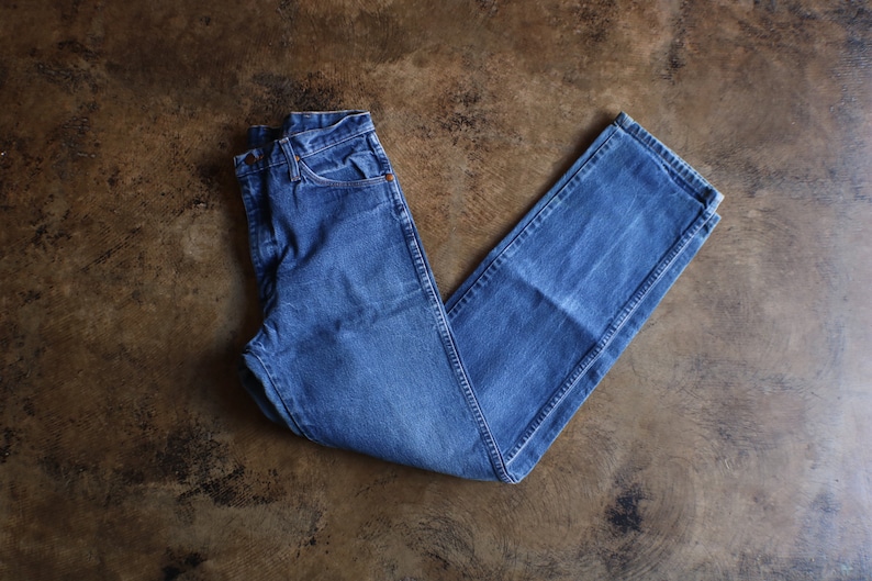 May include: A pair of blue denim jeans, laid flat on a brown surface. The jeans have a classic five-pocket design with a button and zipper closure. The denim has a faded appearance.