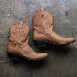 Könnte beinhalten: Ein Paar wadenhohe Cowboystiefel aus hellbraunem Leder mit spitzer Zehenpartie und schwarzen Blockabsätzen. Die Schäfte der Stiefel sind mit tonalen Stickmustern verziert, und das Leder weist durchgehend eine glatte, matte Oberfläche auf.
