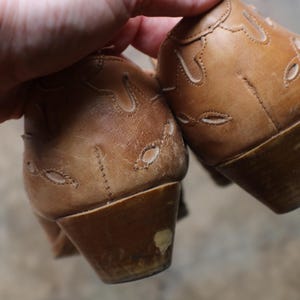 May include: A pair of light brown leather shoes with decorative cut-out patterns. The shoes have a low heel and show signs of wear. The image focuses on the back of the shoes, highlighting the stitching and design details.