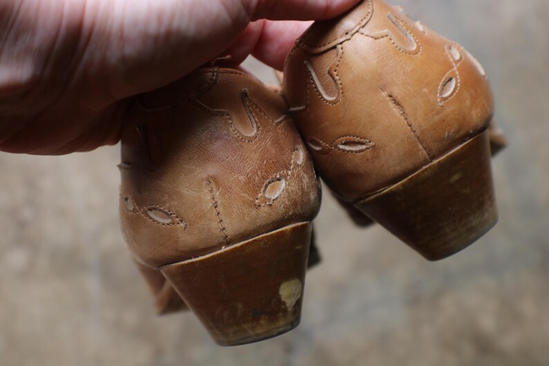 May include: A pair of light brown leather shoes with decorative cut-out patterns. The shoes have a low heel and show signs of wear. The image focuses on the back of the shoes, highlighting the stitching and design details.