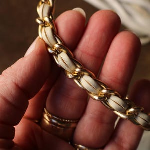 May include: Close-up of a gold chain strap with white leather woven through the links. The strap is held in a hand, with gold rings on the fingers. The chain is a bright gold color.