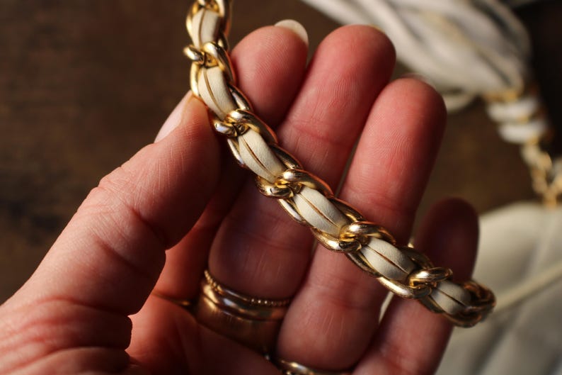 May include: Close-up of a gold chain strap with white leather woven through the links. The strap is held in a hand, with gold rings on the fingers. The chain is a bright gold color.