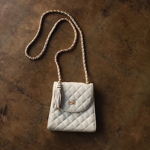 May include: Ivory quilted crossbody bag with a gold chain strap and tassel detail. The bag features a gold bow clasp and a flap closure. The bag is set against a brown, textured background.
