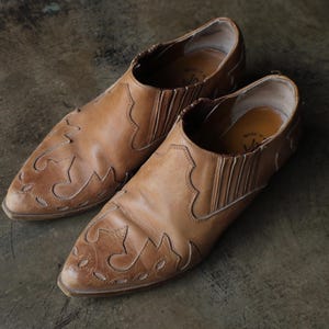 May include: A pair of brown leather slip-on shoes with decorative stitching and pointed toes. The shoes have a worn, vintage appearance, suggesting they are well-loved and have been worn. The leather has a natural, aged look.