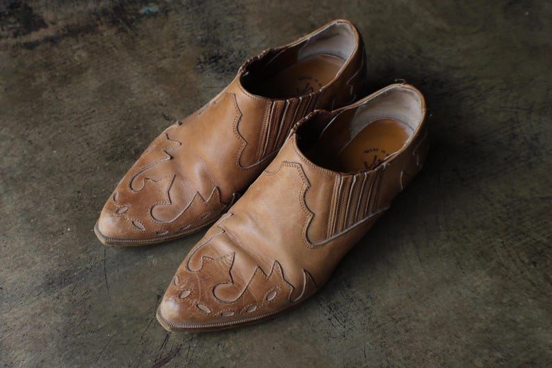 May include: A pair of brown leather slip-on shoes with decorative stitching and pointed toes. The shoes have a worn, vintage appearance, suggesting they are well-loved and have been worn. The leather has a natural, aged look.
