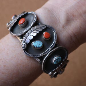 May include: Silver cuff bracelet featuring three oval panels with decorative elements. Each panel has a dark background and is adorned with a coral or turquoise stone. The bracelet has a vintage, handcrafted aesthetic.