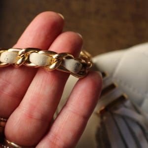 May include: Close-up of a white quilted handbag with a gold chain strap. The chain is interwoven with a cream-colored material. The bag has a tassel detail and gold hardware. The bag is a classic style.