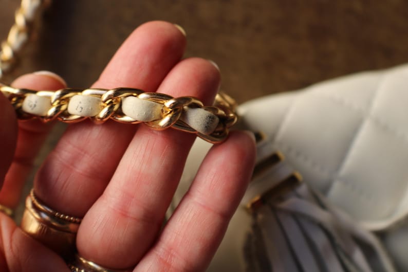 May include: Close-up of a white quilted handbag with a gold chain strap. The chain is interwoven with a cream-colored material. The bag has a tassel detail and gold hardware. The bag is a classic style.