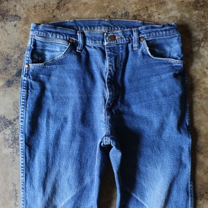 Puede incluir: Primer plano de unos vaqueros azules. Los vaqueros tienen un dise&ntilde;o cl&aacute;sico de cinco bolsillos con remaches de color cobre y cierre de bot&oacute;n. La mezclilla tiene un aspecto descolorido, lo que sugiere desgaste y lavado. Los vaqueros est&aacute;n planos sobre una superficie texturizada.