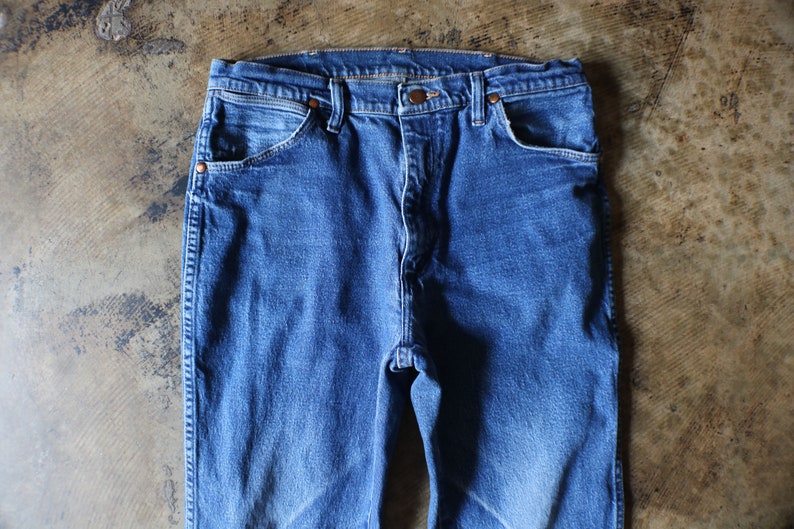 Puede incluir: Primer plano de unos vaqueros azules. Los vaqueros tienen un dise&ntilde;o cl&aacute;sico de cinco bolsillos con remaches de color cobre y cierre de bot&oacute;n. La mezclilla tiene un aspecto descolorido, lo que sugiere desgaste y lavado. Los vaqueros est&aacute;n planos sobre una superficie texturizada.
