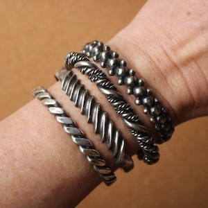 May include: A set of four silver-toned metal cuff bracelets worn together. The stack includes one twisted rope design, one diagonal ribbed band, one braided cable style, and one textured band featuring a series of small, raised spherical beads.