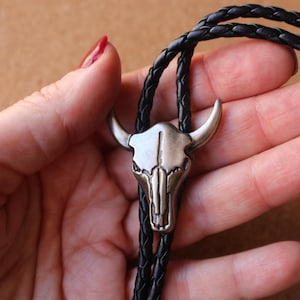 May include: A bolo tie featuring a silver-toned metal steer skull pendant with prominent horns. The pendant is attached to a black braided leather cord that loops through the back of the skull, creating a secure and adjustable neck accessory.