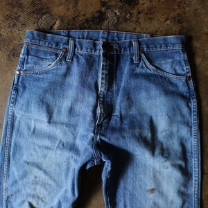 Puede incluir: Primer plano de unos vaqueros azules. Los vaqueros tienen un dise&ntilde;o cl&aacute;sico de cinco bolsillos, con cierre de bot&oacute;n y cremallera. El denim tiene un aspecto descolorido con algo de decoloraci&oacute;n y desgaste.