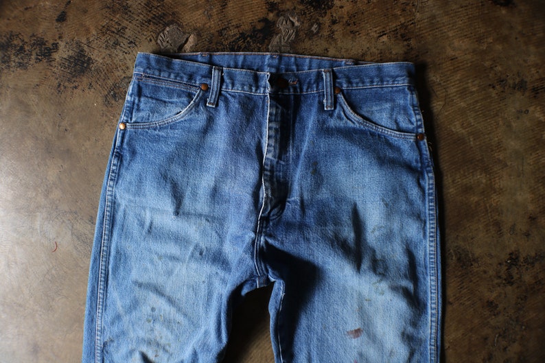 Puede incluir: Primer plano de unos vaqueros azules. Los vaqueros tienen un dise&ntilde;o cl&aacute;sico de cinco bolsillos, con cierre de bot&oacute;n y cremallera. El denim tiene un aspecto descolorido con algo de decoloraci&oacute;n y desgaste.