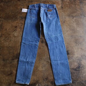 May include: Blue denim jeans with a classic five-pocket design. The jeans feature a brown leather patch and a small tag. The jeans are laid flat on a brown surface.