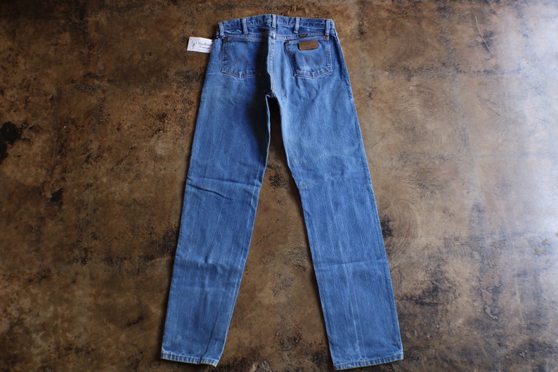 May include: Blue denim jeans with a classic five-pocket design. The jeans feature a brown leather patch and a small tag. The jeans are laid flat on a brown surface.