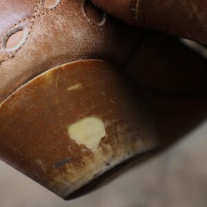 May include: Close-up of a brown leather shoe with a rounded heel. The shoe has decorative cutouts and visible stitching. The heel shows signs of wear with a lighter-colored patch.