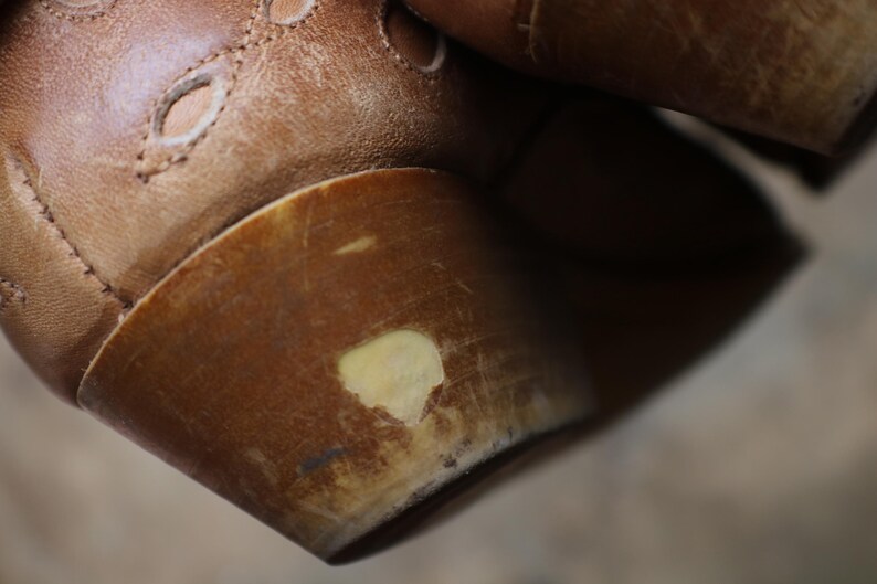 May include: Close-up of a brown leather shoe with a rounded heel. The shoe has decorative cutouts and visible stitching. The heel shows signs of wear with a lighter-colored patch.