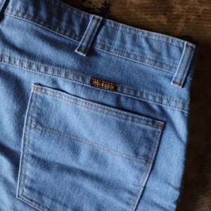 May include: Close-up of light blue denim jeans with a back pocket and the Wrangler brand label. The jeans have tan stitching and belt loops. The fabric appears to be slightly faded, suggesting a vintage style.