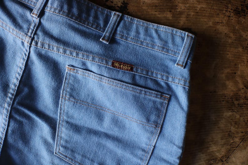 May include: Close-up of light blue denim jeans with a back pocket and the Wrangler brand label. The jeans have tan stitching and belt loops. The fabric appears to be slightly faded, suggesting a vintage style.