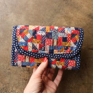 May include: A rectangular quilted wallet featuring a patchwork pattern of small squares in red, blue, yellow, and white floral and geometric prints. The wallet has a dark blue flap with white polka dots and a matching dark blue polka dot border.