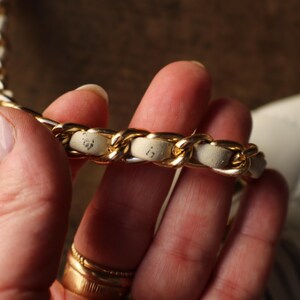May include: Close-up of a gold chain strap with cream-colored leather woven through the links. The strap is part of a white quilted handbag, held by a person. The gold chain is thick and shiny.