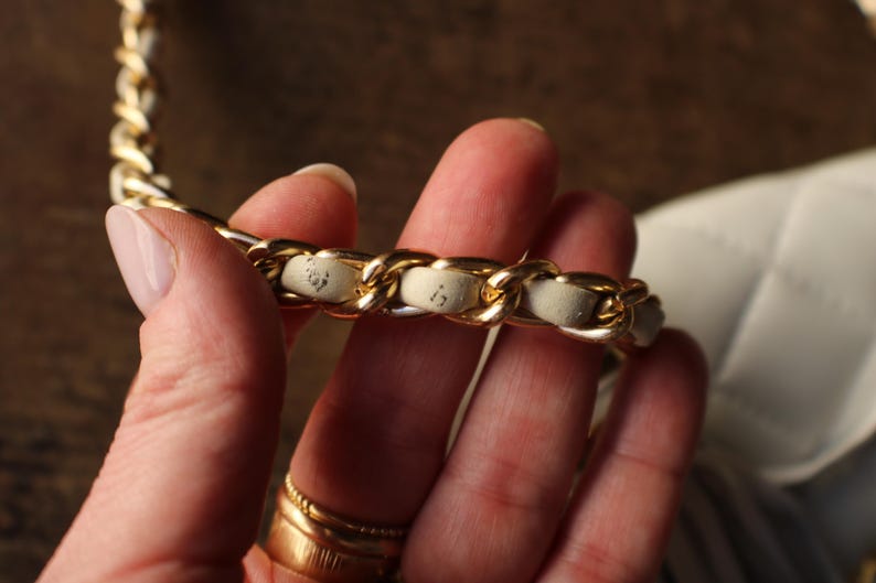 May include: Close-up of a gold chain strap with cream-colored leather woven through the links. The strap is part of a white quilted handbag, held by a person. The gold chain is thick and shiny.
