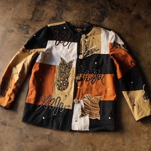 May include: A long-sleeve jacket featuring a patchwork design in black, tan, and white. The garment is decorated with embroidered squiggly lines, animal print fabric patches, and small metallic studs. It closes with three round, patterned buttons.