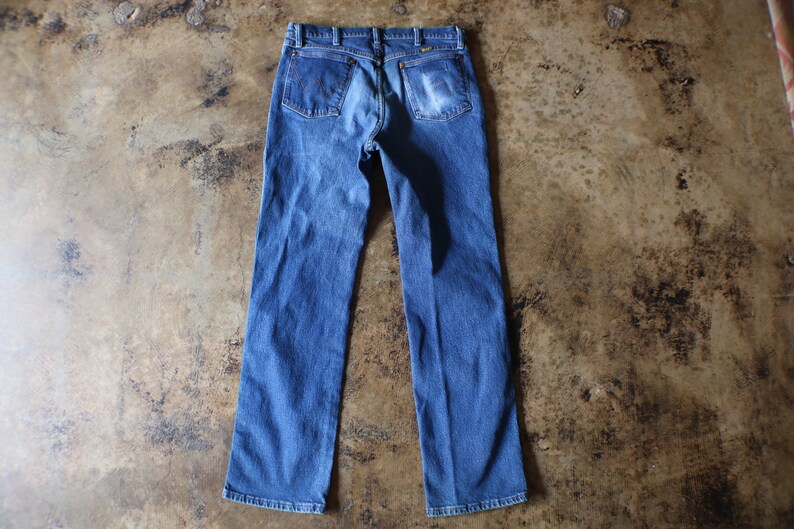 Puede incluir: Vaqueros azules de mezclilla con un dise&ntilde;o cl&aacute;sico de cinco bolsillos. Los vaqueros tienen un lavado descolorido y un corte de pierna recta. Los bolsillos traseros son visibles. Prenda de vestir vintage.