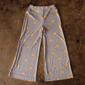 Può includere: Pantaloni a vita alta e gamba larga caratterizzati da un piccolo motivo a scacchi bianchi e neri. Il tessuto è decorato con un motivo ripetitivo di piccole forme circolari giallo brillante, simili a soli, distribuite uniformemente sul capo.
