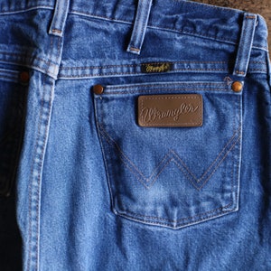 May include: Close-up of blue denim Wrangler jeans. The jeans feature a brown leather patch with the Wrangler logo on the back pocket. The stitching is a contrasting brown color. The jeans have a classic five-pocket design.