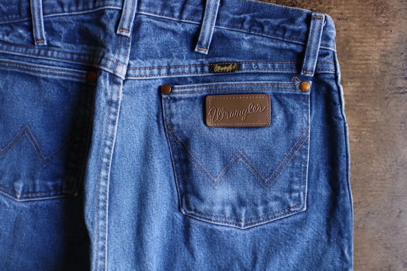 May include: Close-up of blue denim Wrangler jeans. The jeans feature a brown leather patch with the Wrangler logo on the back pocket. The stitching is a contrasting brown color. The jeans have a classic five-pocket design.