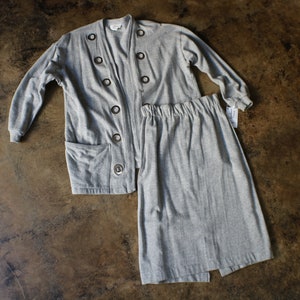May include: A gray two-piece knit set, including a long-sleeved cardigan with large eyelet button closures and a matching skirt. The skirt has an elastic waistband. The set is displayed on a brown surface.