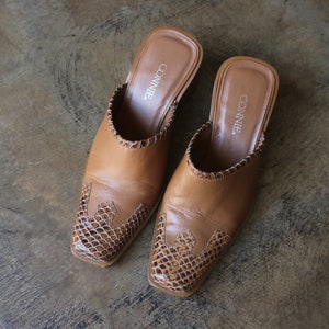 May include: A pair of tan leather slip-on mules with a square toe and a snakeskin pattern on the vamp. The shoes have a stitched trim around the opening and the brand name "CONNIE" is visible inside. The shoes are on a neutral surface.