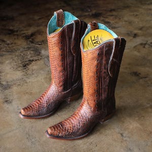May include: A pair of brown cowboy boots with a snakeskin pattern. The boots have a pointed toe, a tall shaft, and a turquoise blue interior. The boots are displayed on a brown surface.