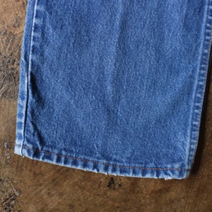 Puede incluir: Primer plano de una secci&oacute;n de vaqueros azules. La tela es de un lavado medio con textura visible. El dobladillo est&aacute; terminado con costuras naranjas. Los vaqueros est&aacute;n sobre una superficie marr&oacute;n texturizada.