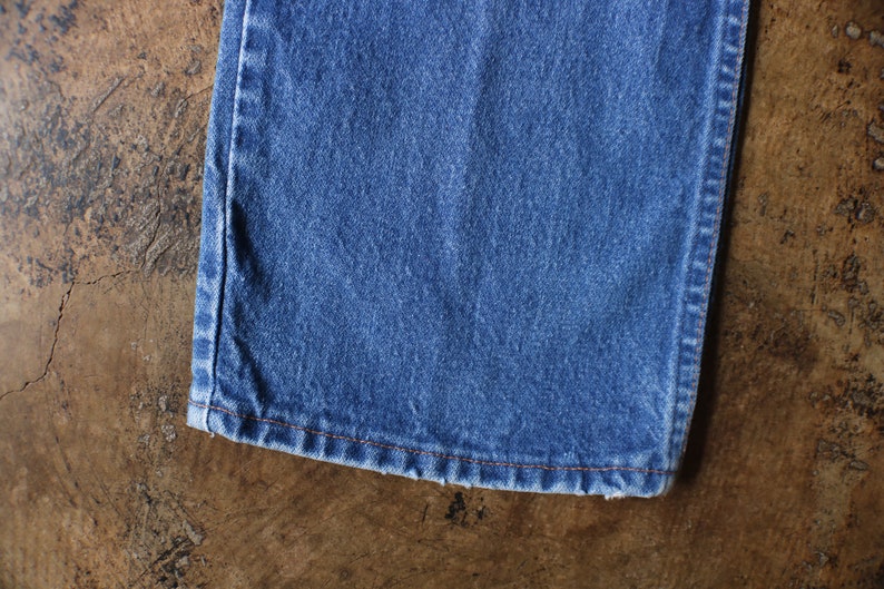 Puede incluir: Primer plano de una secci&oacute;n de vaqueros azules. La tela es de un lavado medio con textura visible. El dobladillo est&aacute; terminado con costuras naranjas. Los vaqueros est&aacute;n sobre una superficie marr&oacute;n texturizada.