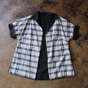 May include: A short-sleeved, open-front shirt with a black collar and cuffs. The shirt features a plaid pattern in cream, black, and navy blue. The garment is laid flat on a brown surface.