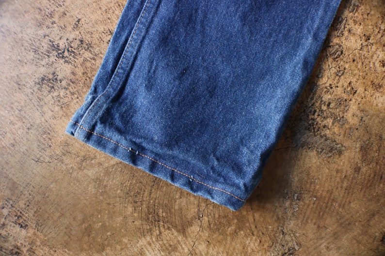 Puede incluir: Primer plano de una secci&oacute;n de vaqueros azules. La tela es de un color &iacute;ndigo intenso, con textura visible y sutiles variaciones de tono. Las costuras naranjas contrastan con la tela. Los vaqueros est&aacute;n doblados.