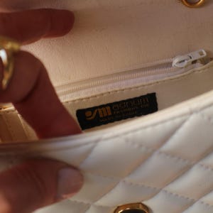 May include: Cream-colored quilted handbag with a gold chain strap. The interior features a zippered pocket and a label that reads "Sanum Fashions Inc. Made in China." The bag is open, revealing the interior lining.