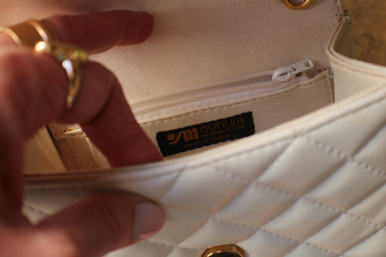 May include: Cream-colored quilted handbag with a gold chain strap. The interior features a zippered pocket and a label that reads "Sanum Fashions Inc. Made in China." The bag is open, revealing the interior lining.