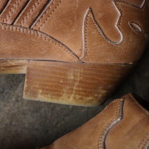 May include: Close-up of a worn, brown leather cowboy boot. The boot features decorative stitching and a stacked heel. The leather shows signs of wear, adding to its rustic appeal. The heel is approximately 2 inches high.