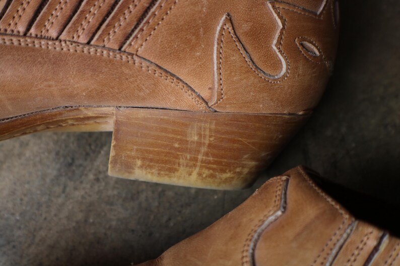 May include: Close-up of a worn, brown leather cowboy boot. The boot features decorative stitching and a stacked heel. The leather shows signs of wear, adding to its rustic appeal. The heel is approximately 2 inches high.